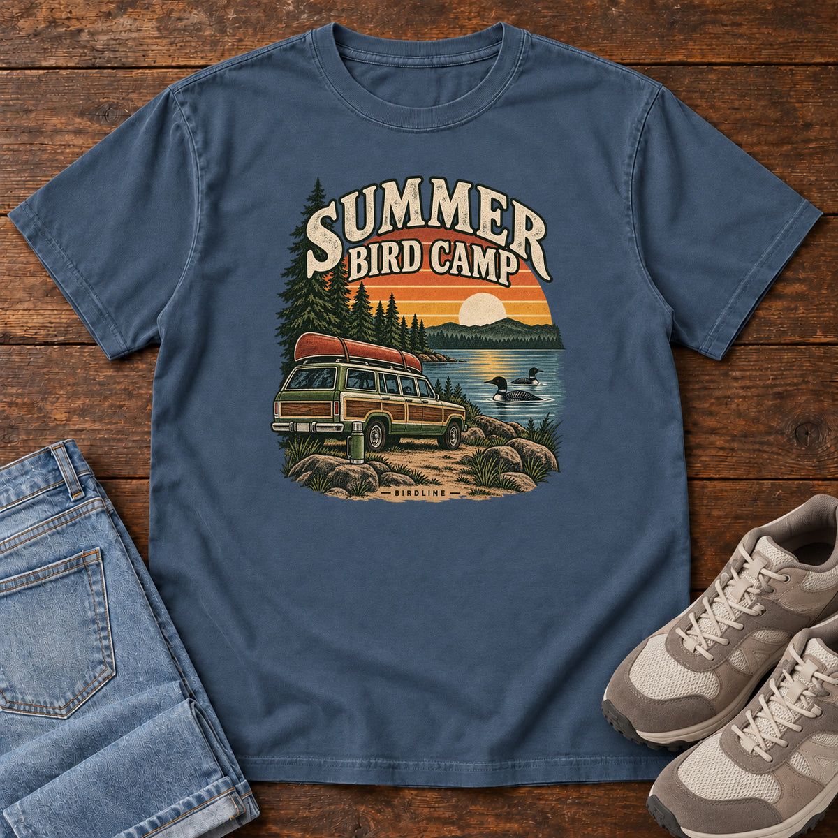 Summer Bird Camp T-Shirt in Midnight, front laid flat on wood, featuring a canoe-topped vehicle beside a lake with swimming birds, pine trees and a striped sunset.