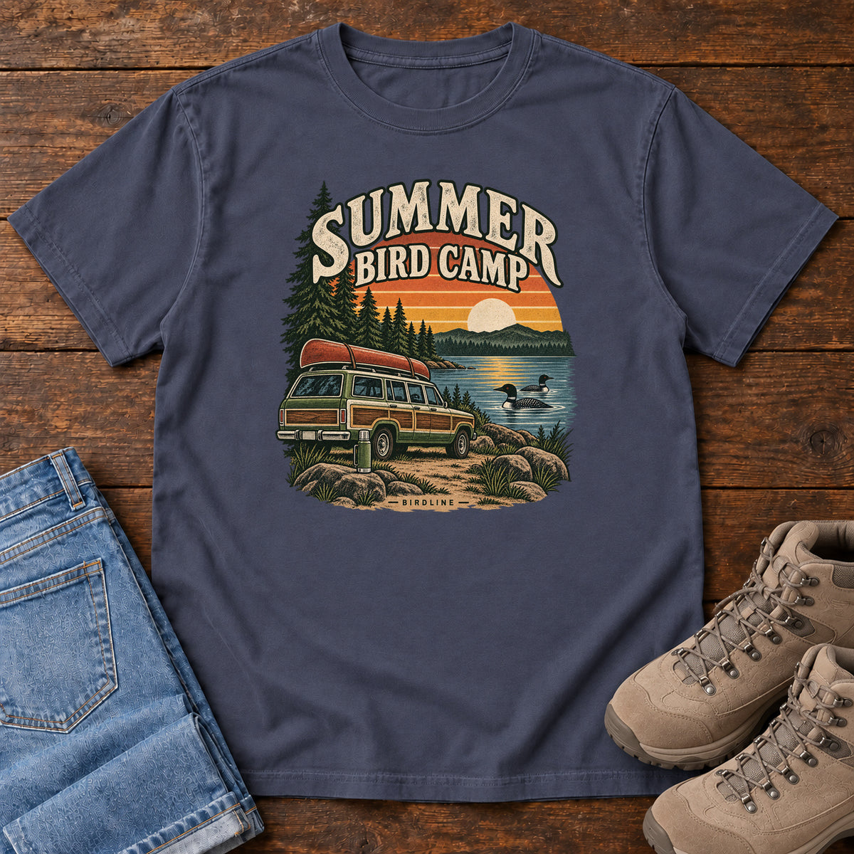 Summer Bird Camp T-Shirt in Navy, front laid flat on wood, featuring a canoe-topped vehicle beside a lake with swimming birds, pine trees and a striped sunset.