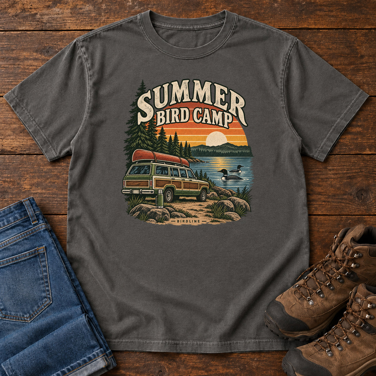 Summer Bird Camp T-Shirt in Pepper, front laid flat on wood, featuring a canoe-topped vehicle beside a lake with swimming birds, pine trees and a striped sunset.