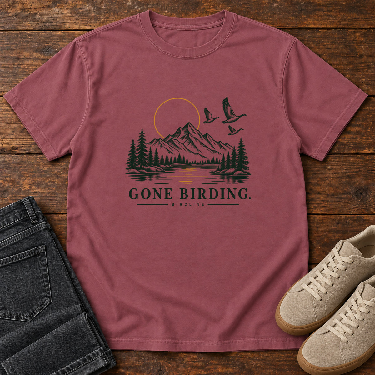 Brick Gone Birding T-Shirt with mountains, evergreen trees, flying birds and an outlined golden sun, front flat lay on wood.