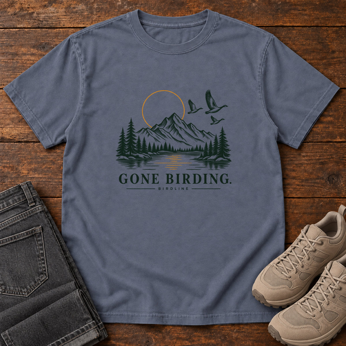 Denim Gone Birding T-Shirt with mountains, evergreen trees, flying birds and an outlined golden sun, front flat lay on wood.