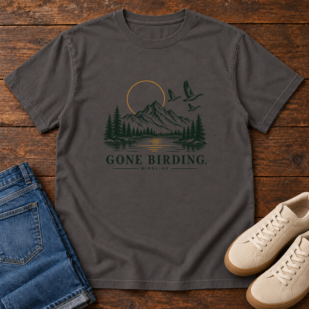 Graphite Gone Birding T-Shirt with mountains, evergreen trees, flying birds and an outlined golden sun, front flat lay on wood.