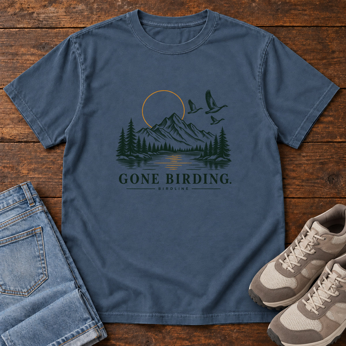 Midnight Gone Birding T-Shirt with mountains, evergreen trees, flying birds and an outlined golden sun, front flat lay on wood.
