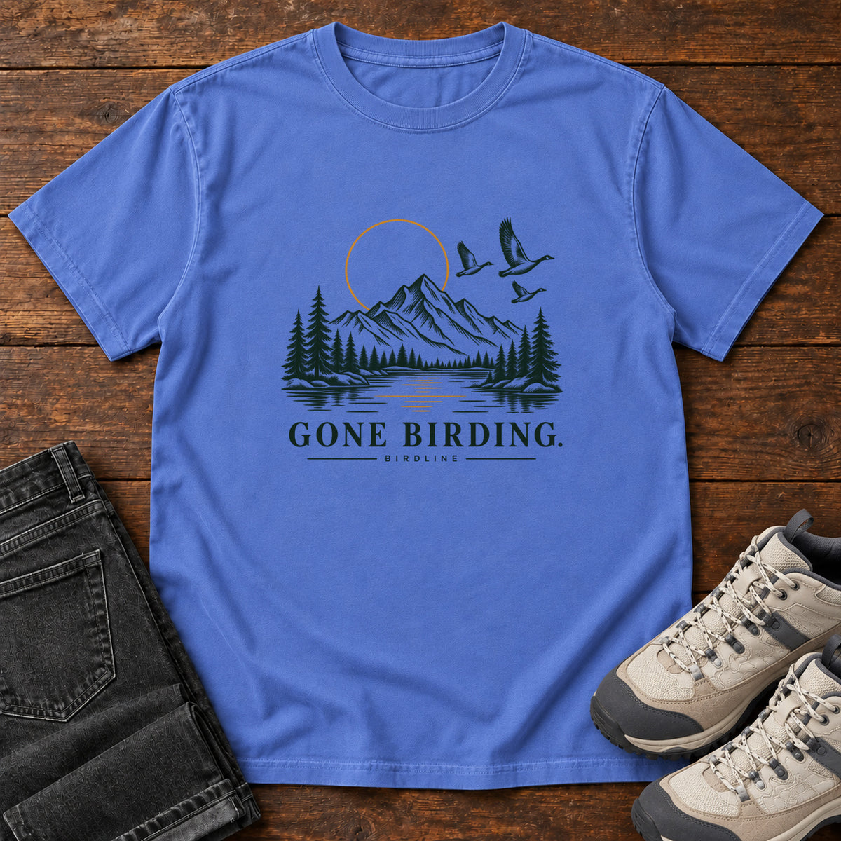 Mystic Blue Gone Birding T-Shirt with mountains, evergreen trees, flying birds and an outlined golden sun, front flat lay on wood.