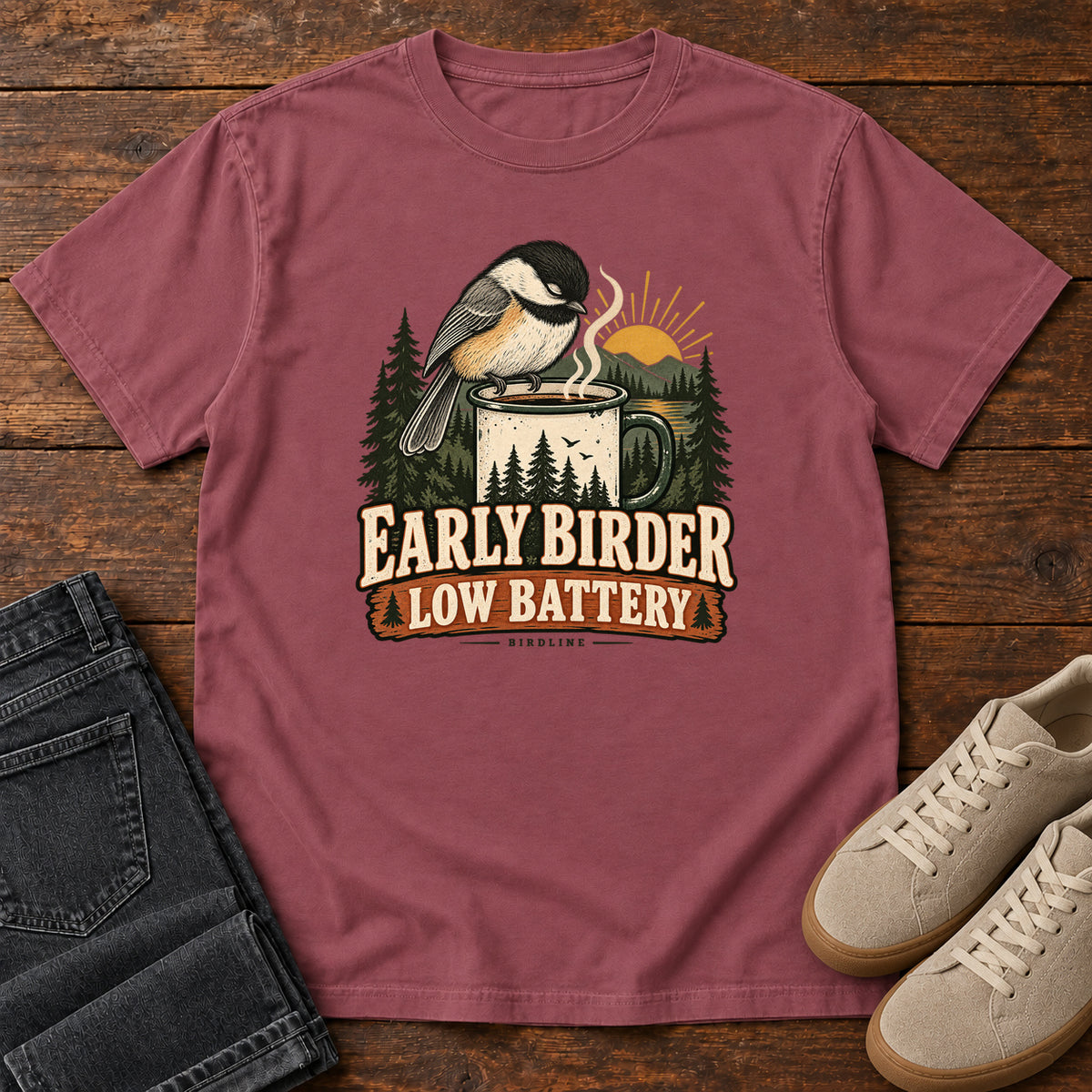 Brick Early Birder. Low Battery. T-Shirt with a bird perched on a steaming mug, evergreens and a rising sun; front flat lay on wood.