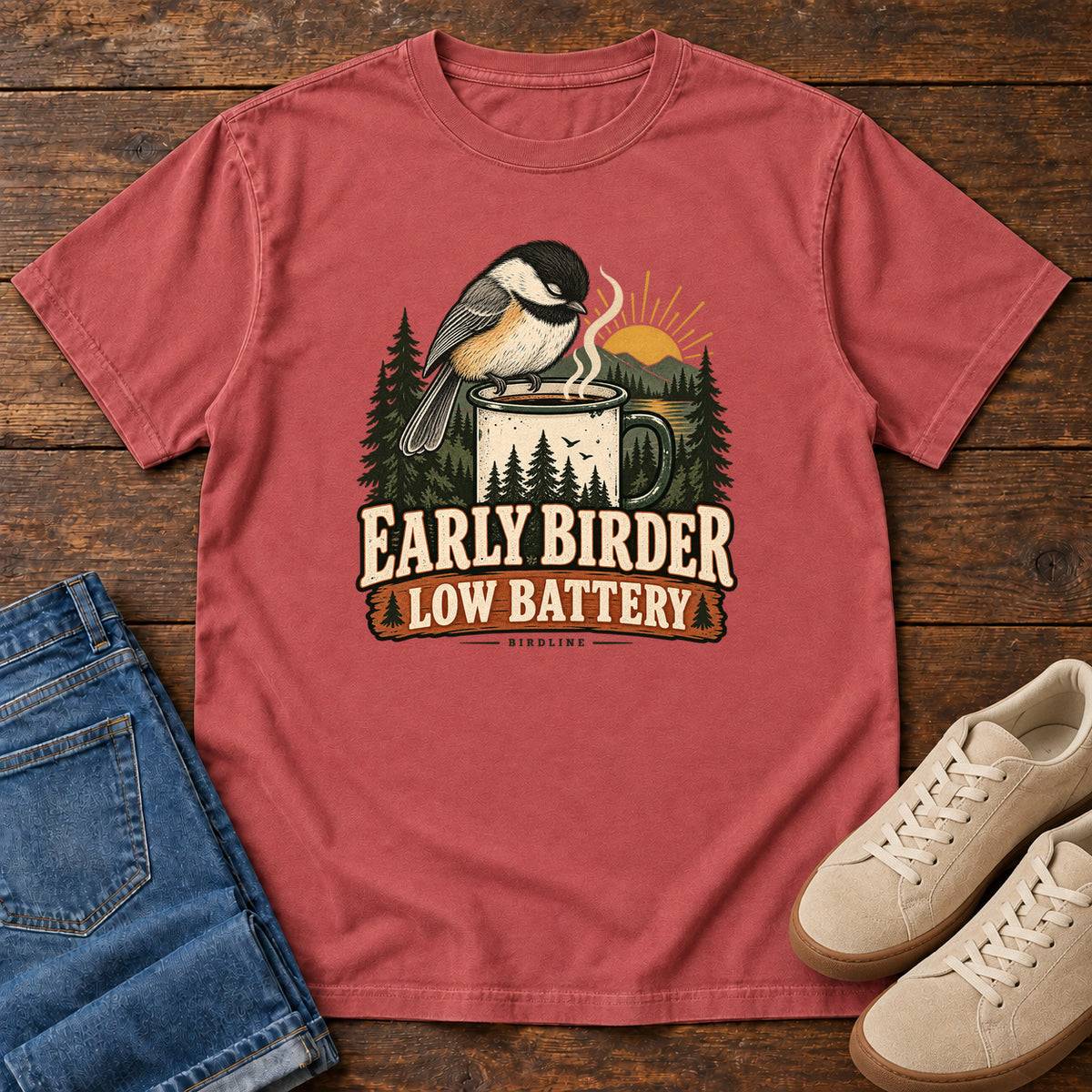 Crimson Early Birder. Low Battery. T-Shirt with a bird perched on a steaming mug, evergreens and a rising sun; front flat lay on wood.