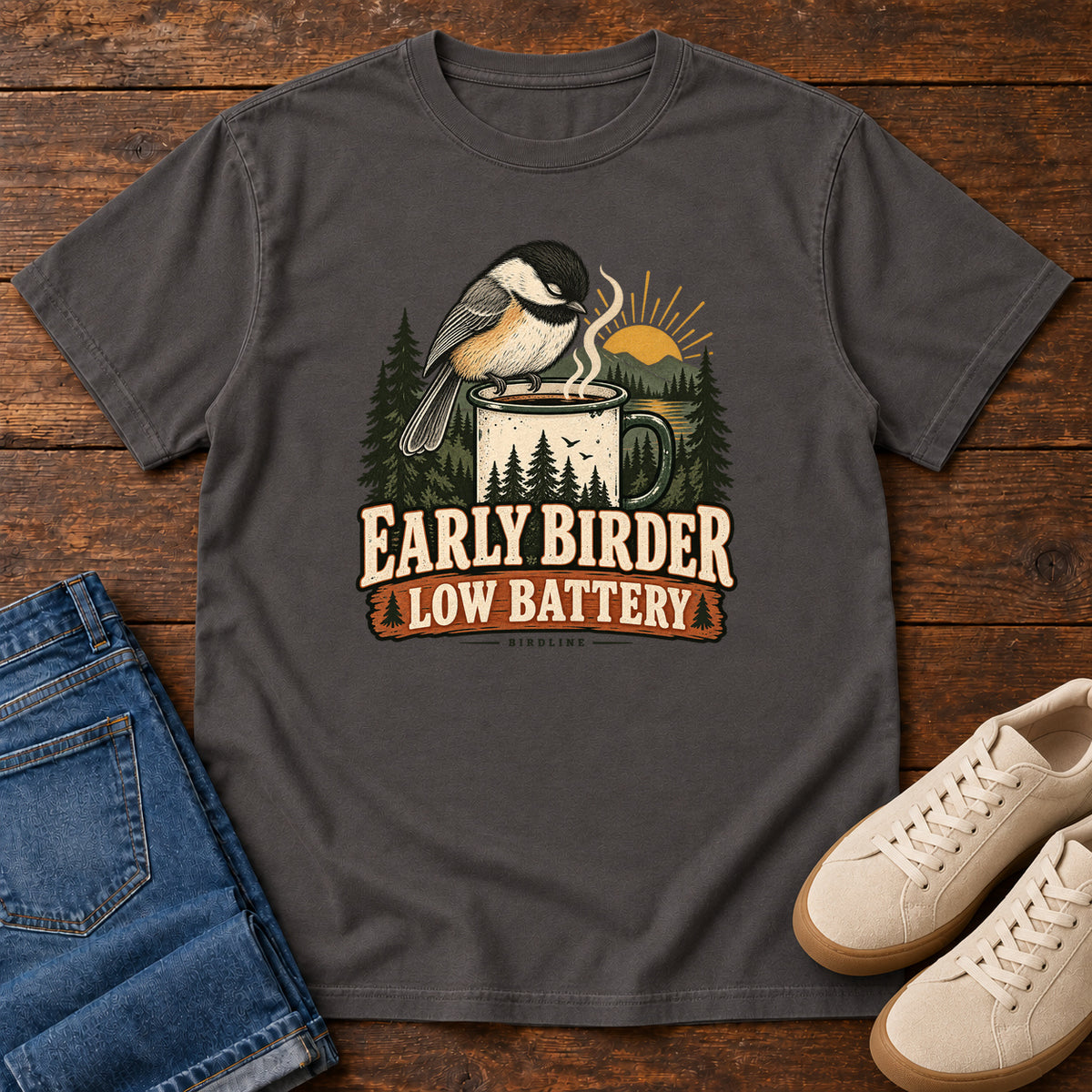 Graphite Early Birder. Low Battery. T-Shirt with a bird perched on a steaming mug, evergreens and a rising sun; front flat lay on wood.