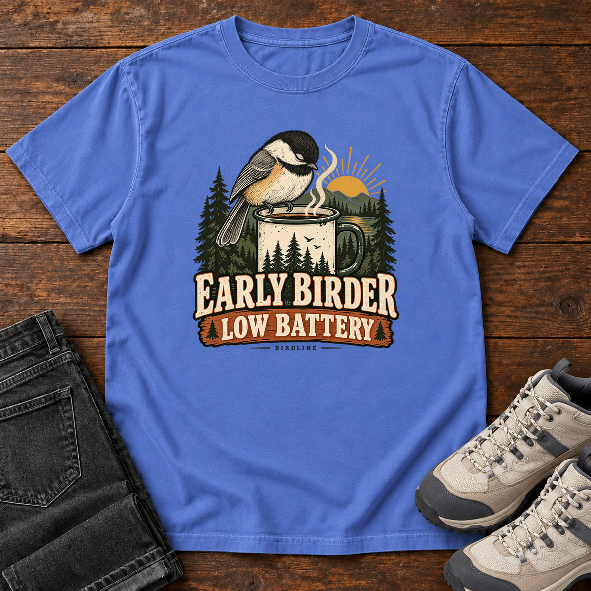 Mystic Blue Early Birder. Low Battery. T-Shirt with a bird perched on a steaming mug, evergreens and a rising sun; front flat lay on wood.