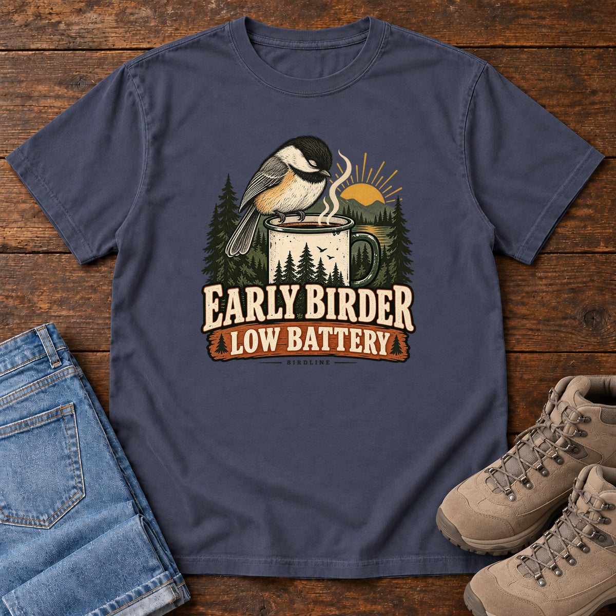Navy Early Birder. Low Battery. T-Shirt with a bird perched on a steaming mug, evergreens and a rising sun; front flat lay on wood.
