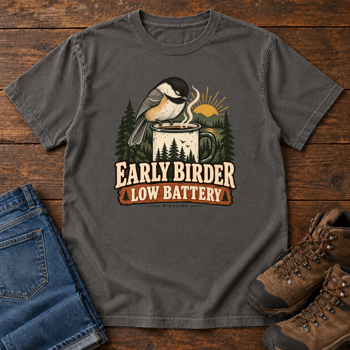 Pepper Early Birder. Low Battery. T-Shirt with a bird perched on a steaming mug, evergreens and a rising sun; front flat lay on wood.