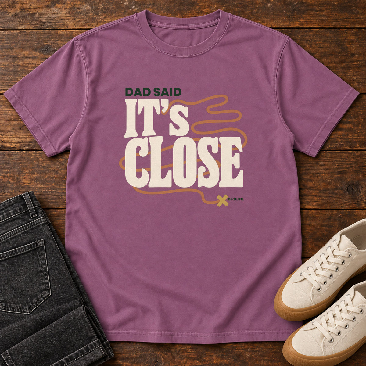 Berry Dad Said It's Close T-Shirt with bold cream lettering wrapped by a winding orange route ending at an X; front flat lay on wood.