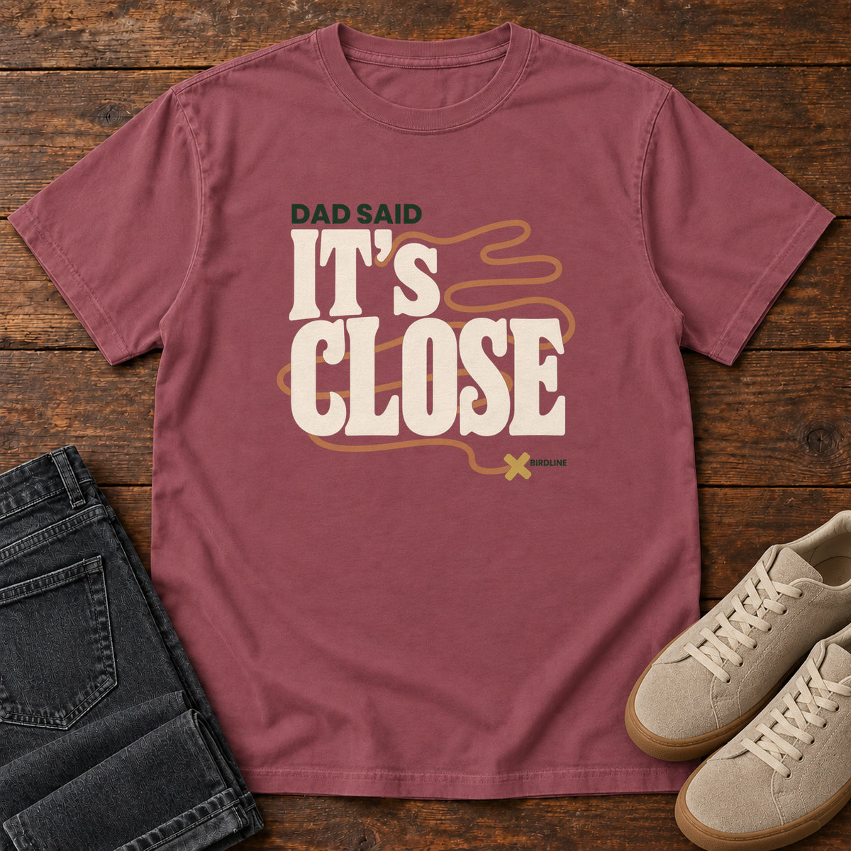 Brick Dad Said It's Close T-Shirt with bold cream lettering wrapped by a winding orange route ending at an X; front flat lay on wood.