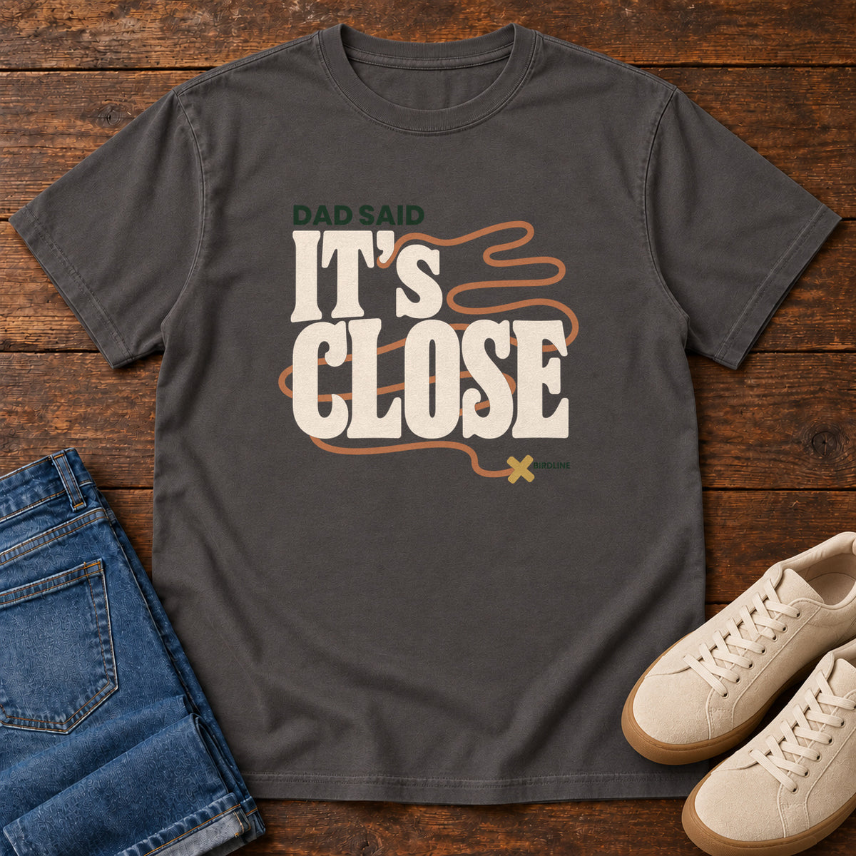 Graphite Dad Said It's Close T-Shirt with bold cream lettering wrapped by a winding orange route ending at an X; front flat lay on wood.