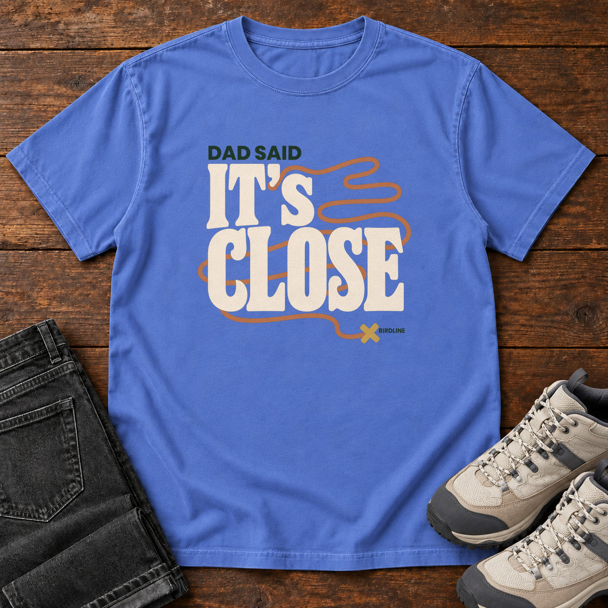 Mystic Blue Dad Said It's Close T-Shirt with bold cream lettering wrapped by a winding orange route ending at an X; front flat lay on wood.