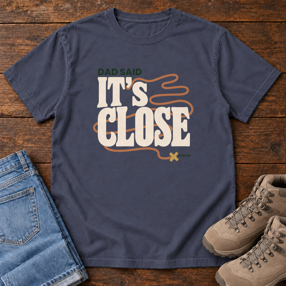 Navy Dad Said It's Close T-Shirt with bold cream lettering wrapped by a winding orange route ending at an X; front flat lay on wood.