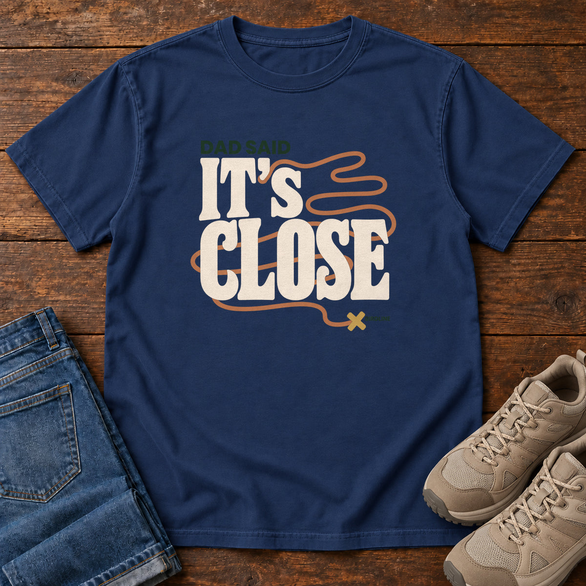 True Navy Dad Said It's Close T-Shirt with bold cream lettering wrapped by a winding orange route ending at an X; front flat lay on wood.