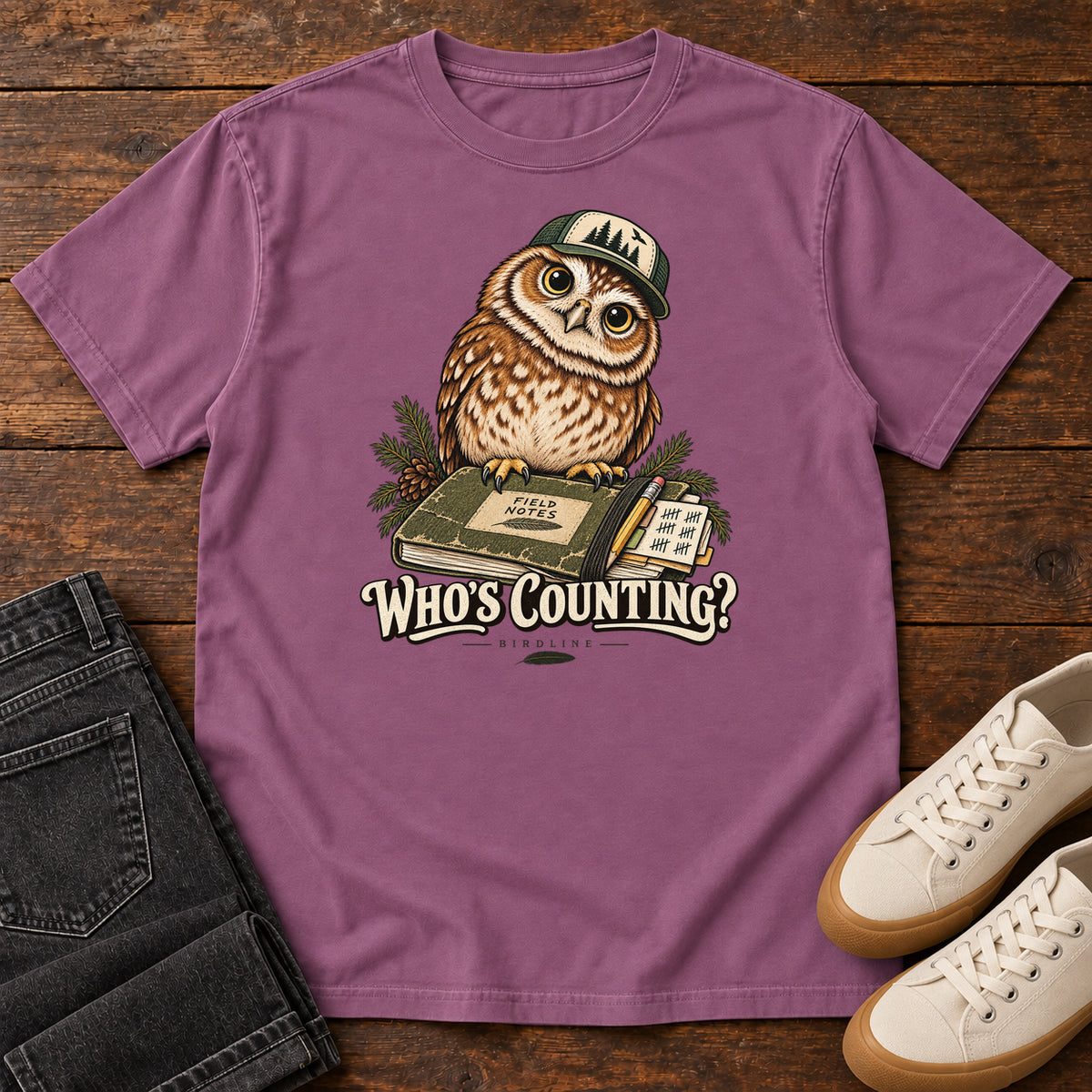 Berry Who's Counting? T-Shirt with a brown owl wearing a green cap beside a field notebook and checklist above cream lettering, front flat lay on wood.