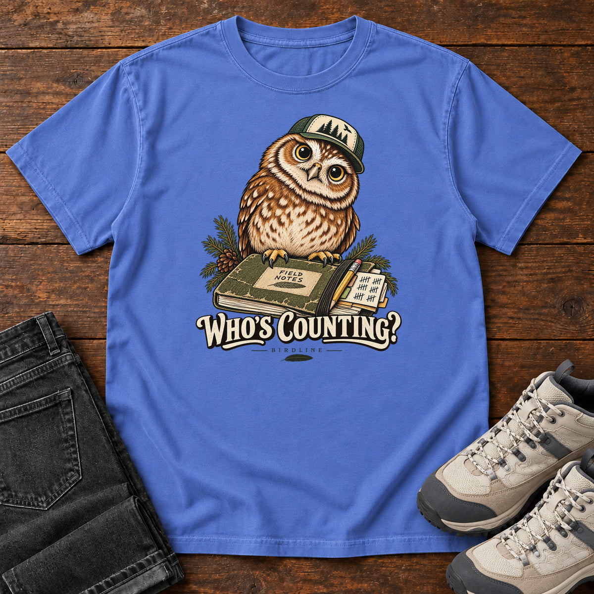 Mystic Blue Who's Counting? T-Shirt with a brown owl wearing a green cap beside a field notebook and checklist above cream lettering, front flat lay on wood.