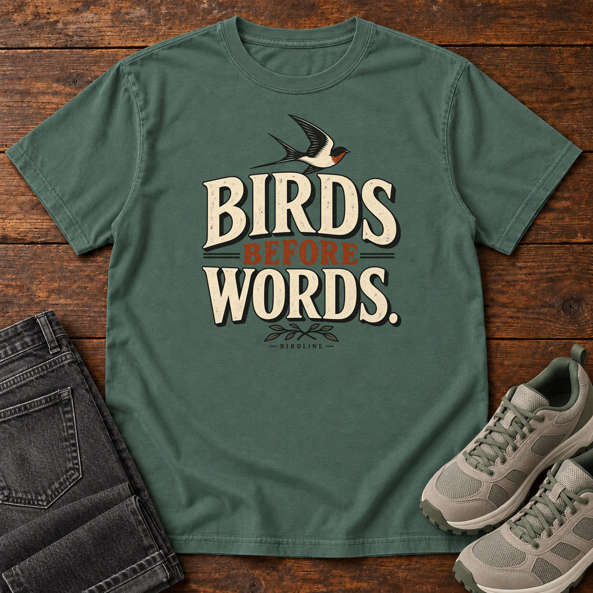 Birds Before Words T-Shirt in Blue Spruce, front laid flat on wood, featuring cream and rust slogan lettering with a small flying bird above and leafy sprigs below.