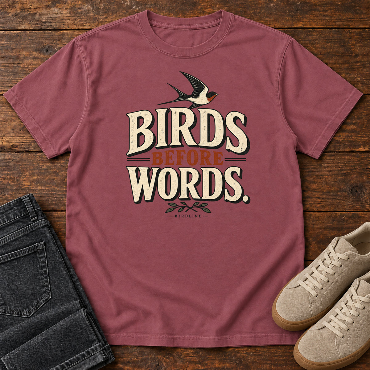 Birds Before Words T-Shirt in Brick, front laid flat on wood, featuring cream and rust slogan lettering with a small flying bird above and leafy sprigs below.