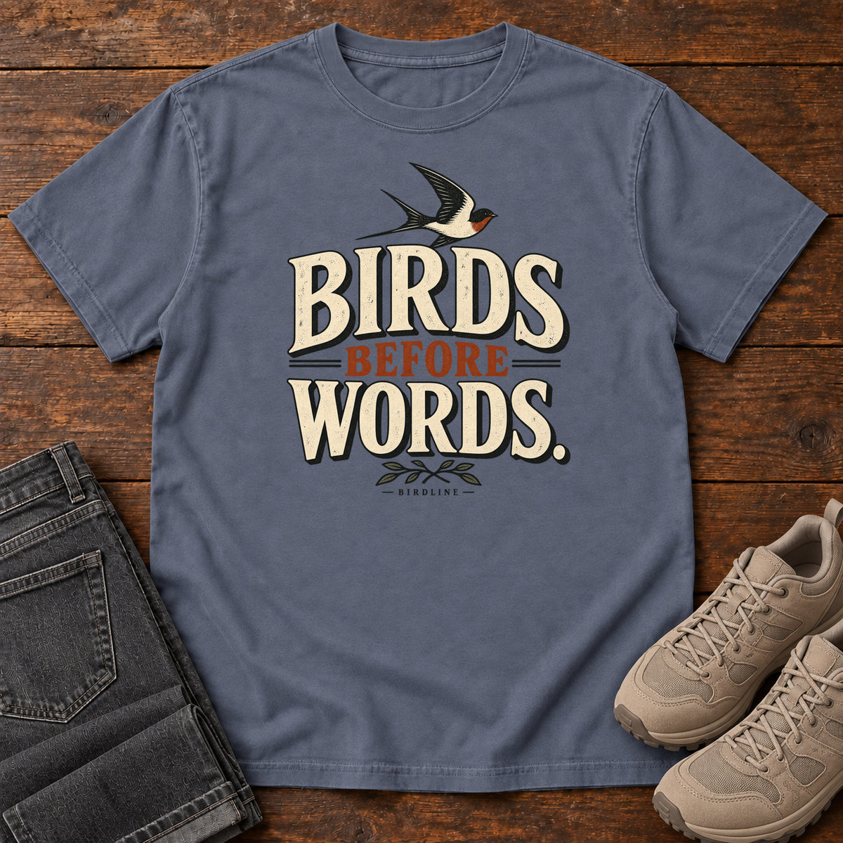 Birds Before Words T-Shirt in Denim, front laid flat on wood, featuring cream and rust slogan lettering with a small flying bird above and leafy sprigs below.