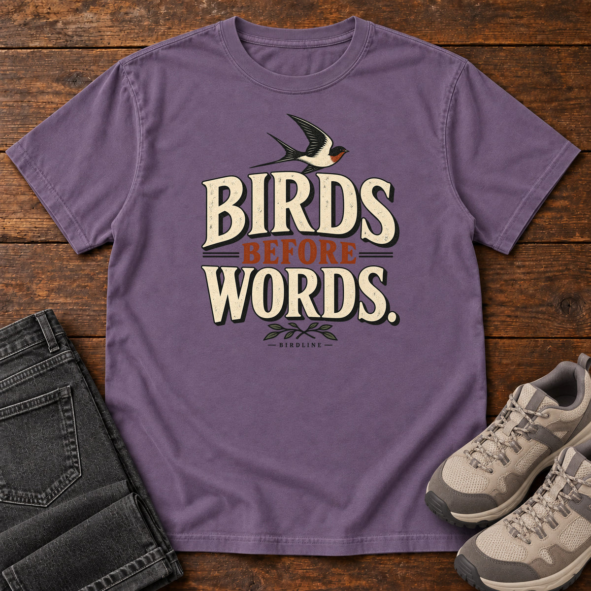 Birds Before Words T-Shirt in Grape, front laid flat on wood, featuring cream and rust slogan lettering with a small flying bird above and leafy sprigs below.