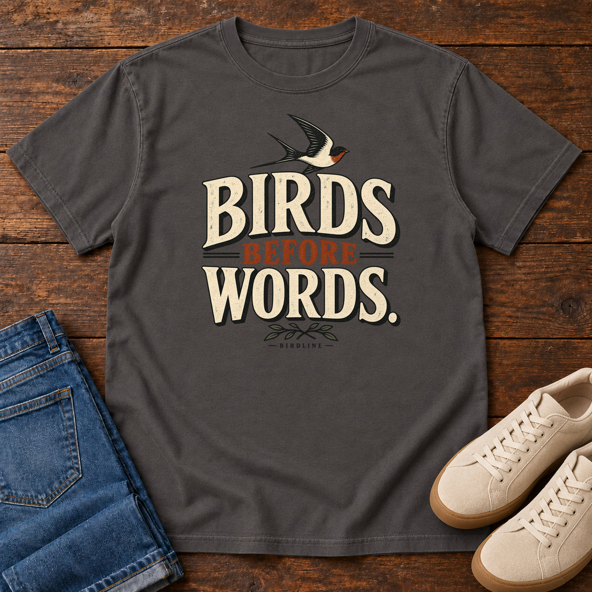 Birds Before Words T-Shirt in Graphite, front laid flat on wood, featuring cream and rust slogan lettering with a small flying bird above and leafy sprigs below.