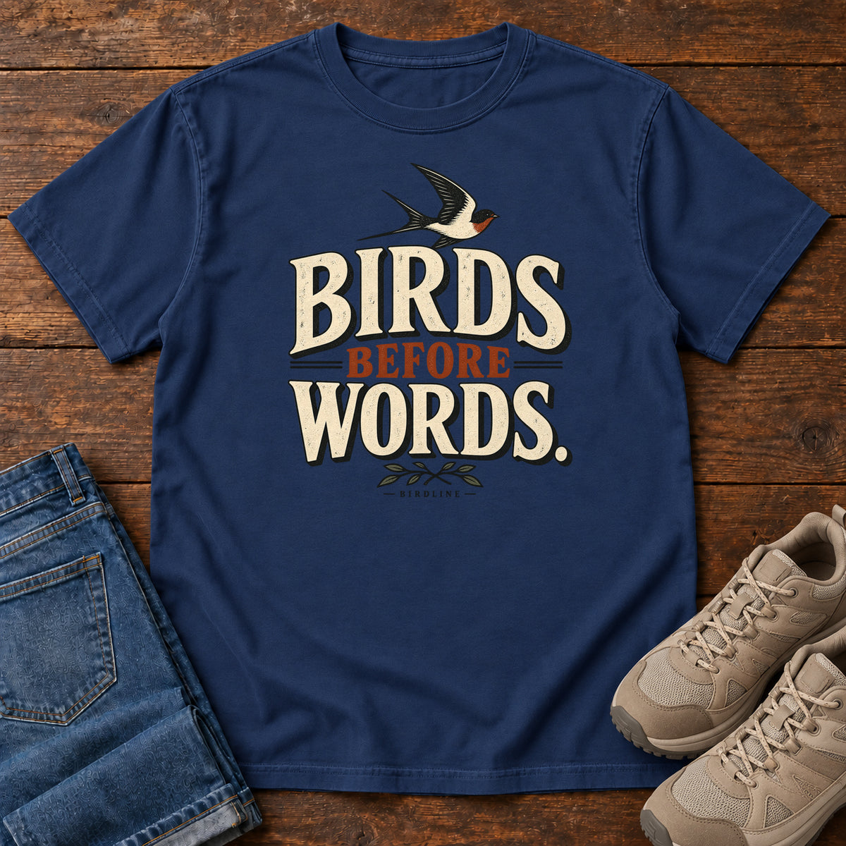 Birds Before Words T-Shirt in True Navy, front laid flat on wood, featuring cream and rust slogan lettering with a small flying bird above and leafy sprigs below.