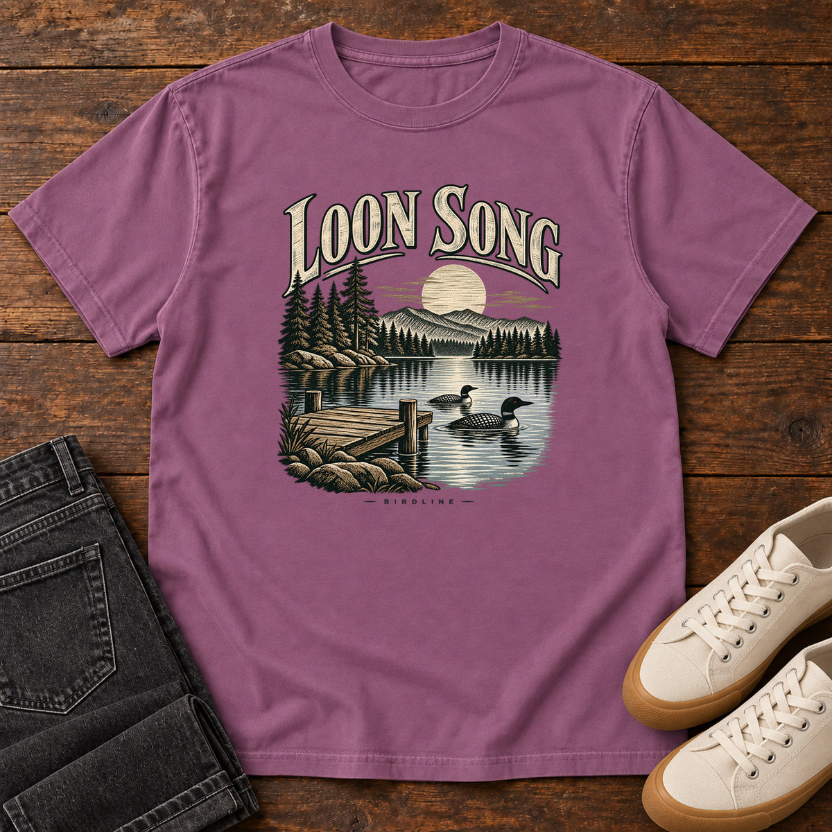 Berry Loon Song T-Shirt with two waterbirds beside a dock on a moonlit lake beneath cream lettering, front flat lay on wood.