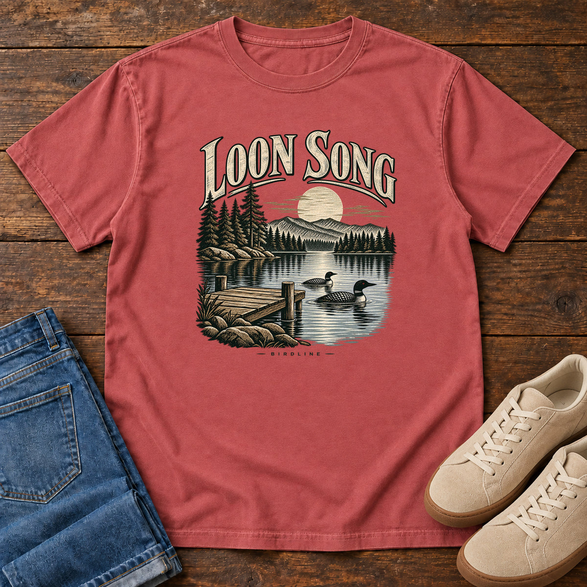 Crimson Loon Song T-Shirt with two waterbirds beside a dock on a moonlit lake beneath cream lettering, front flat lay on wood.