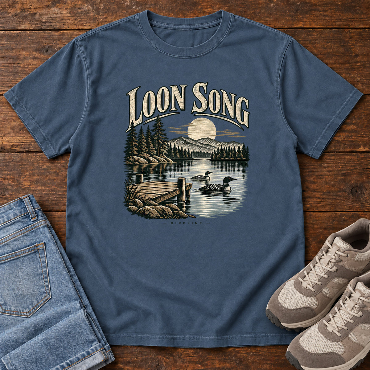 Midnight Loon Song T-Shirt with two waterbirds beside a dock on a moonlit lake beneath cream lettering, front flat lay on wood.