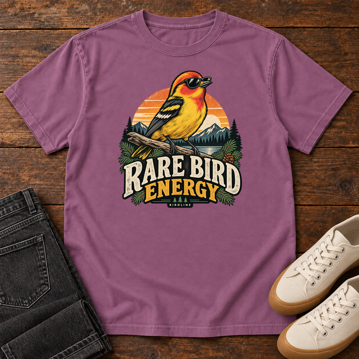 Berry Rare Bird Energy T-Shirt with a yellow and orange bird wearing sunglasses before a striped sunset; front flat lay on wood.