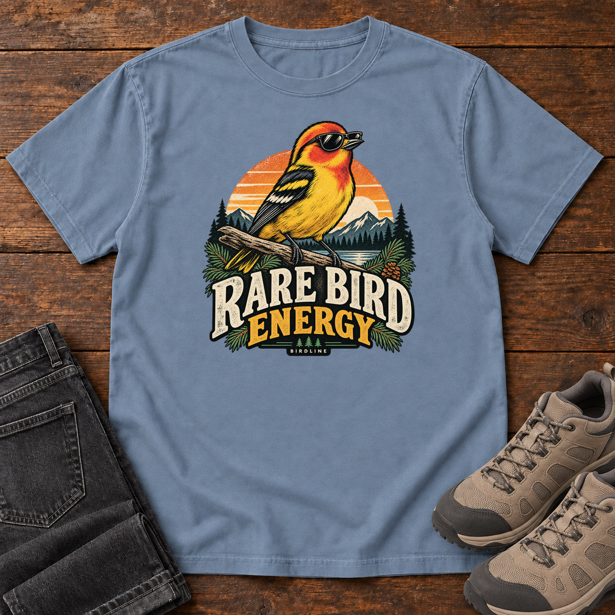 Blue Jean Rare Bird Energy T-Shirt with a yellow and orange bird wearing sunglasses before a striped sunset; front flat lay on wood.