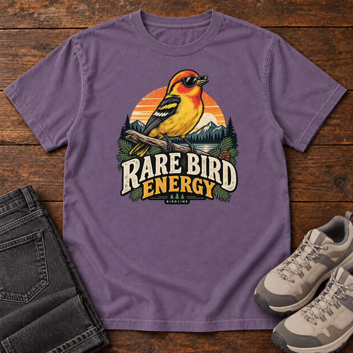 Grape Rare Bird Energy T-Shirt with a yellow and orange bird wearing sunglasses before a striped sunset; front flat lay on wood.
