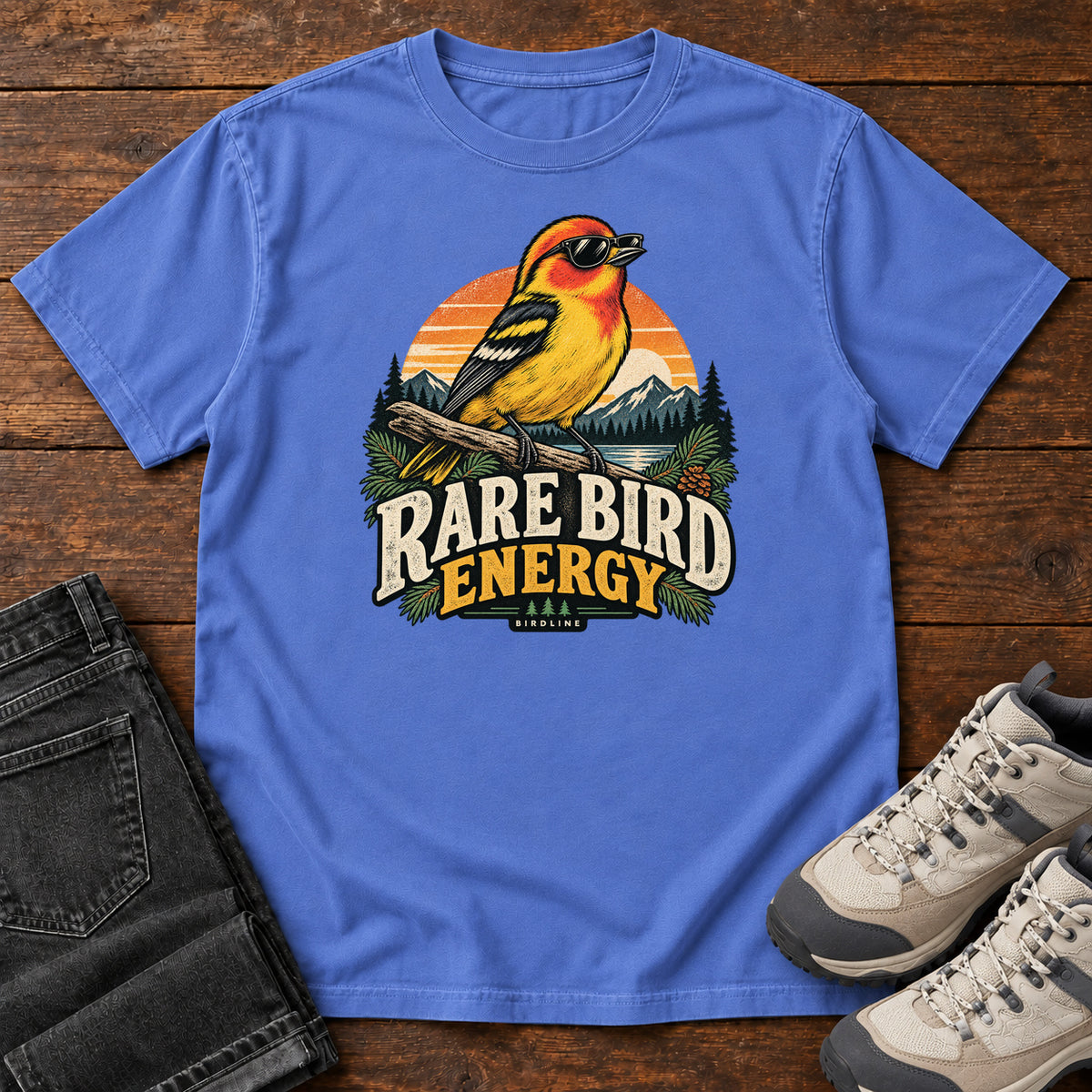 Mystic Blue Rare Bird Energy T-Shirt with a yellow and orange bird wearing sunglasses before a striped sunset; front flat lay on wood.