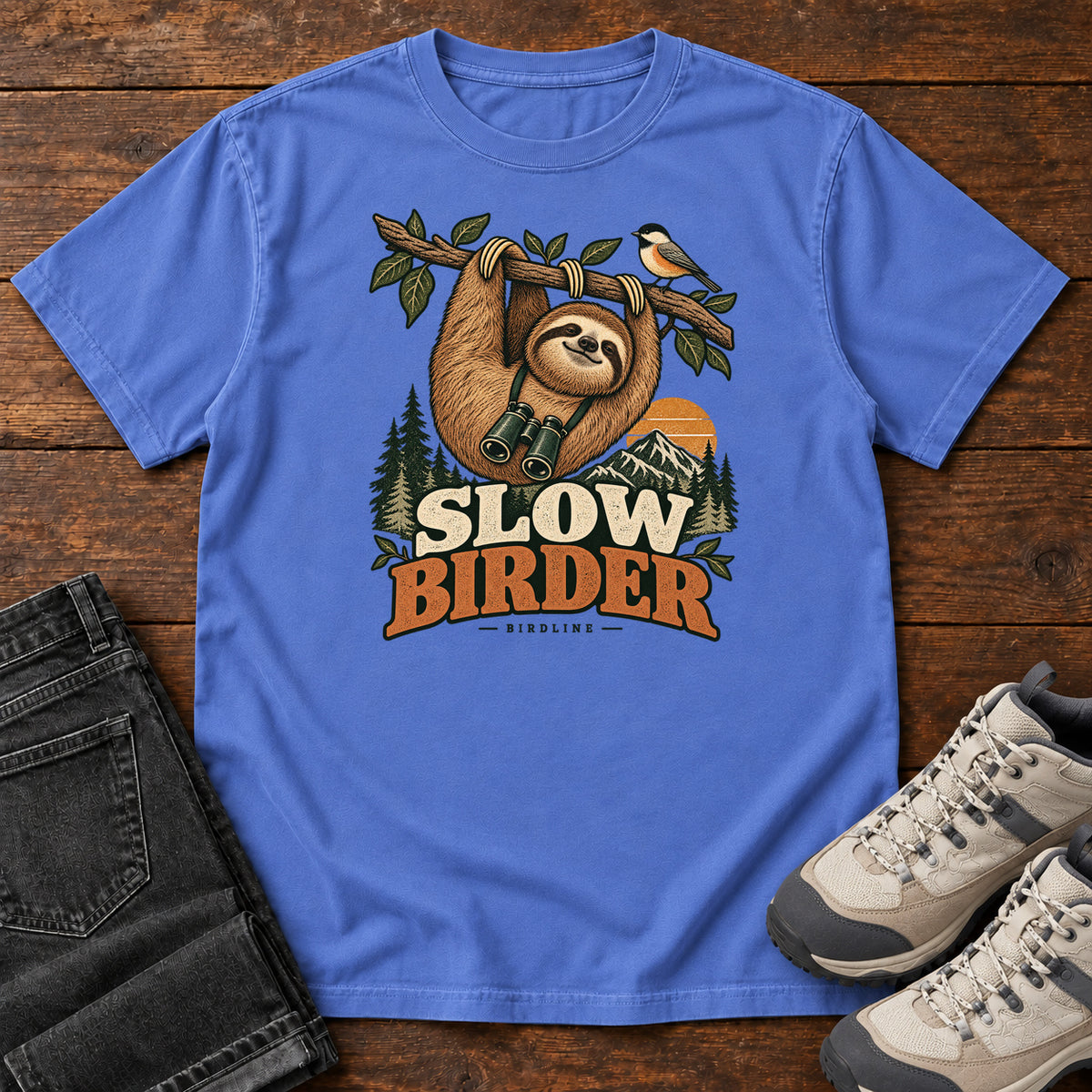 Mystic Blue Slow Birder T-Shirt with a sloth with binoculars hanging from a branch beside a bird and mountains; front flat lay on wood.