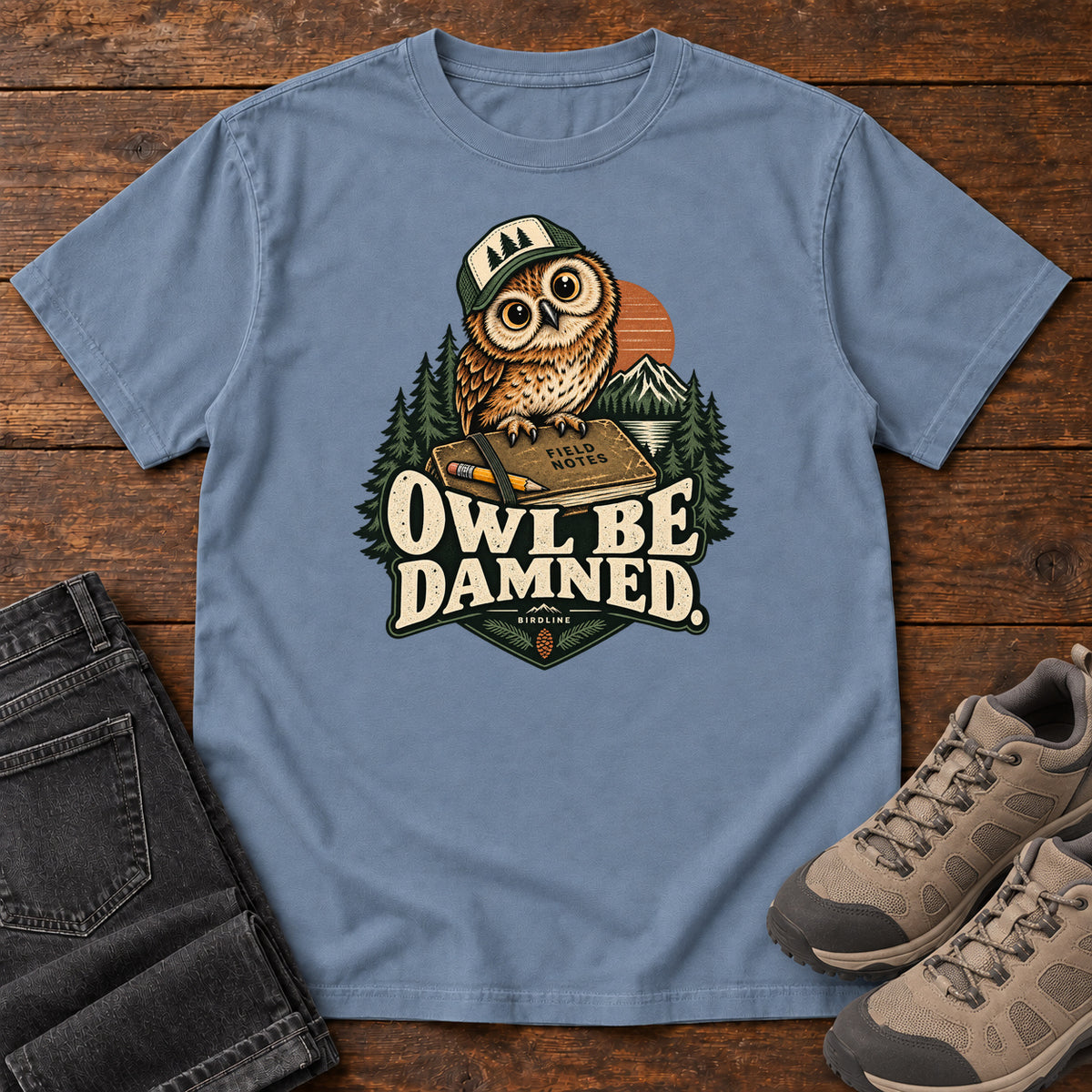 Blue Jean Owl Be Damned. T-Shirt with a brown owl wearing a green cap on a field notebook, with mountains, trees and cream lettering, front flat lay on wood.