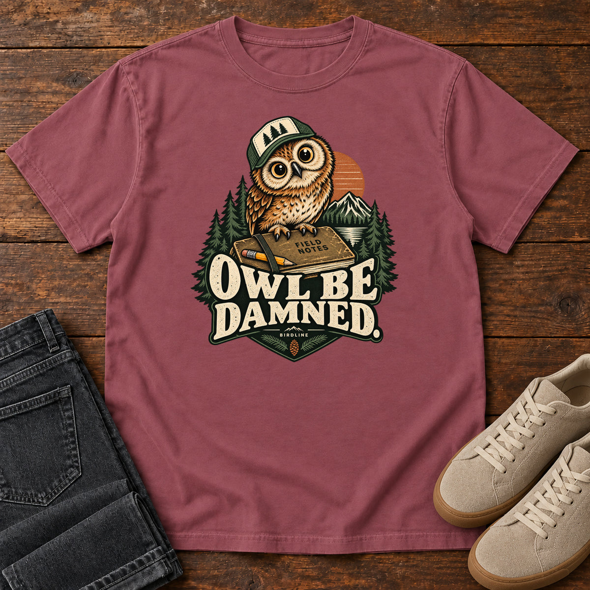 Brick Owl Be Damned. T-Shirt with a brown owl wearing a green cap on a field notebook, with mountains, trees and cream lettering, front flat lay on wood.