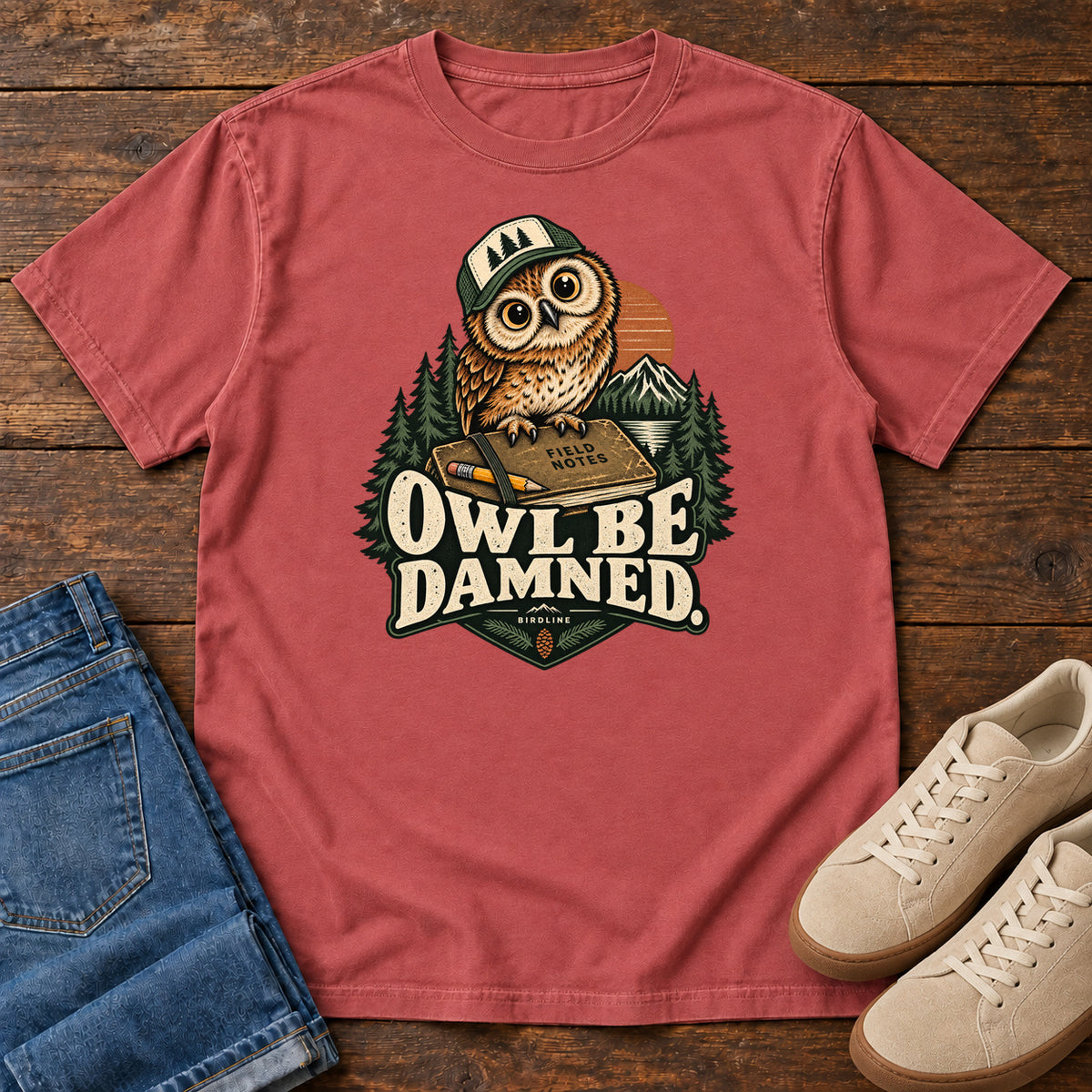 Crimson Owl Be Damned. T-Shirt with a brown owl wearing a green cap on a field notebook, with mountains, trees and cream lettering, front flat lay on wood.