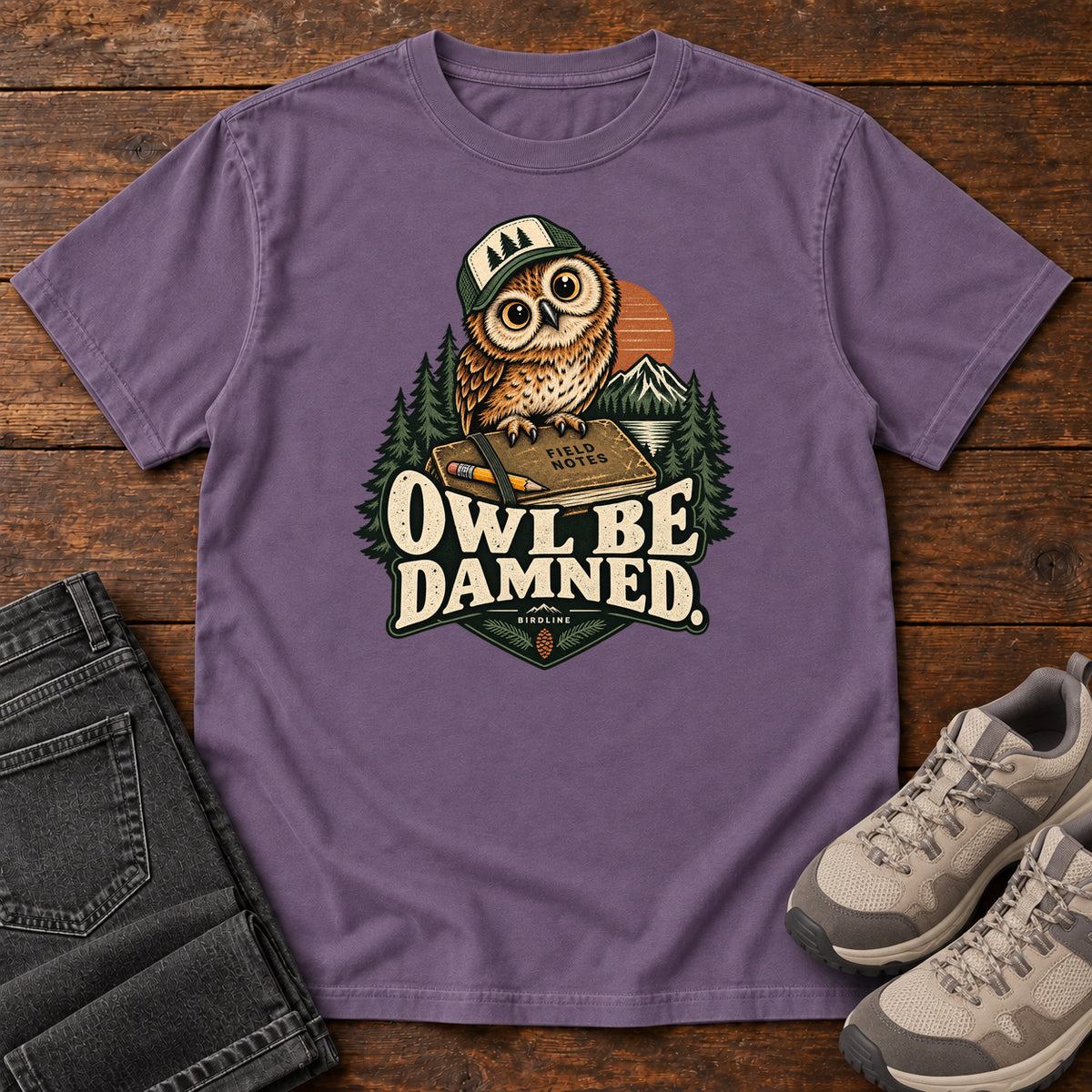 Grape Owl Be Damned. T-Shirt with a brown owl wearing a green cap on a field notebook, with mountains, trees and cream lettering, front flat lay on wood.