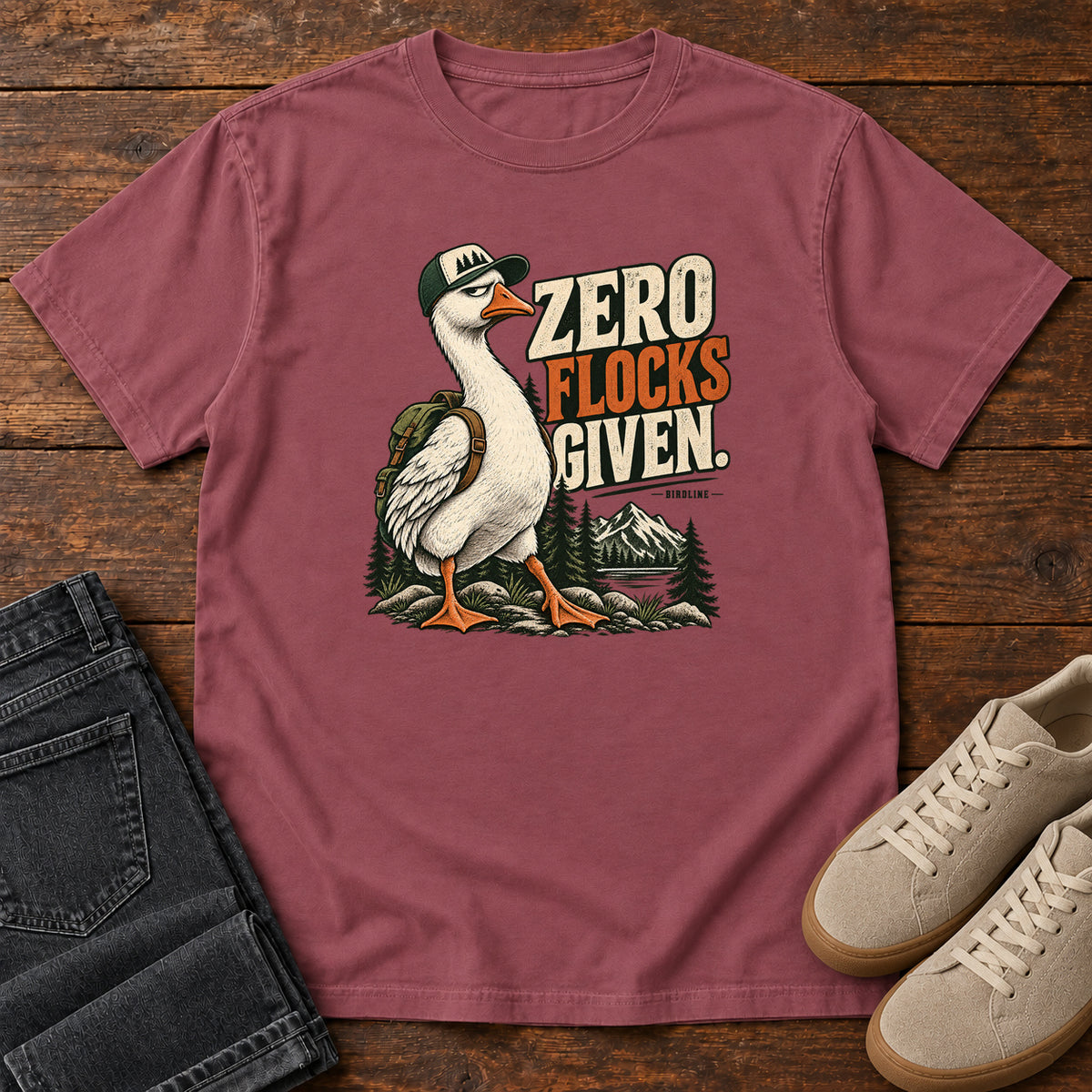 Zero Flocks Given T-Shirt in Brick, front laid flat on wood, featuring a white long-necked bird wearing a cap and backpack beside cream-and-orange lettering and mountains.