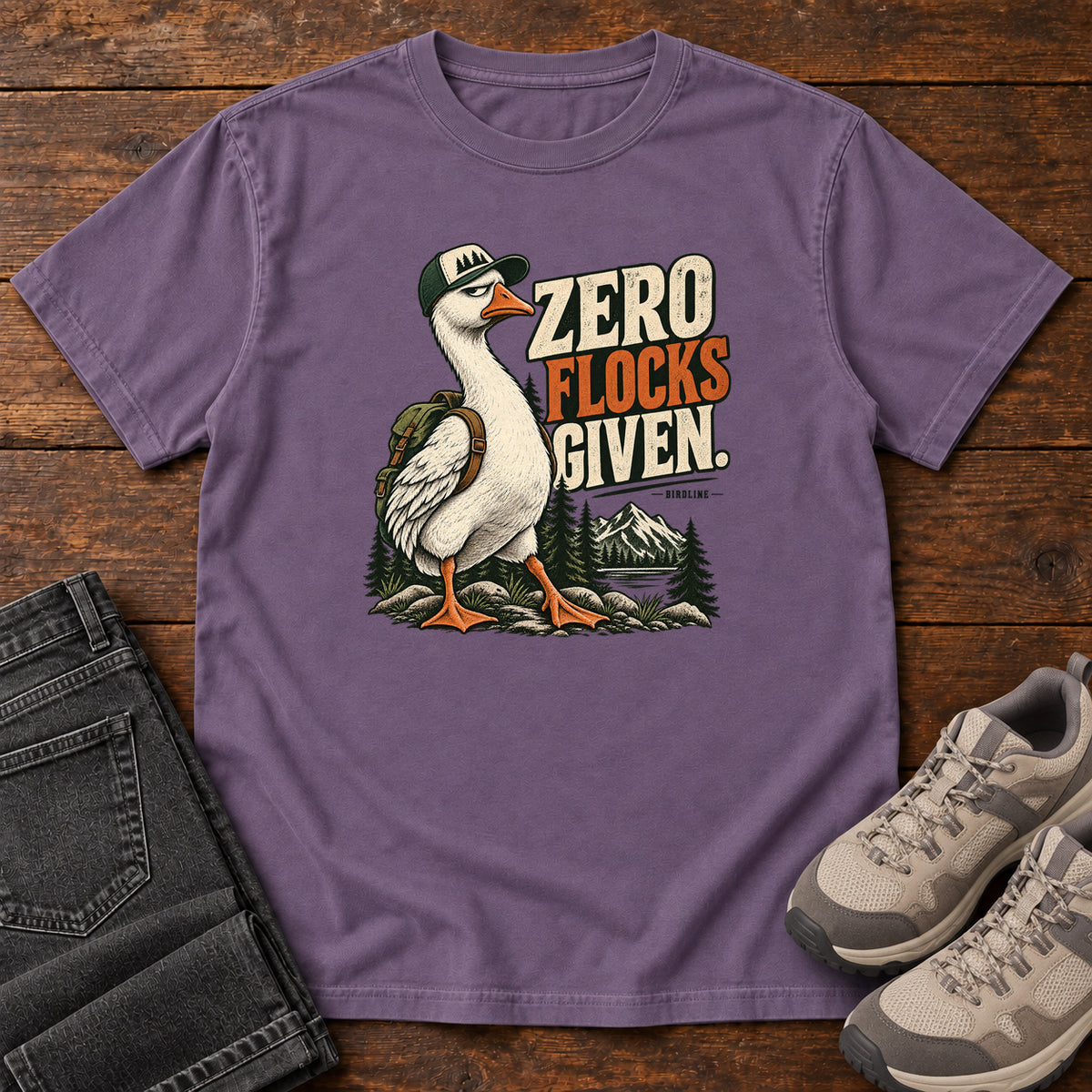 Zero Flocks Given T-Shirt in Grape, front laid flat on wood, featuring a white long-necked bird wearing a cap and backpack beside cream-and-orange lettering and mountains.