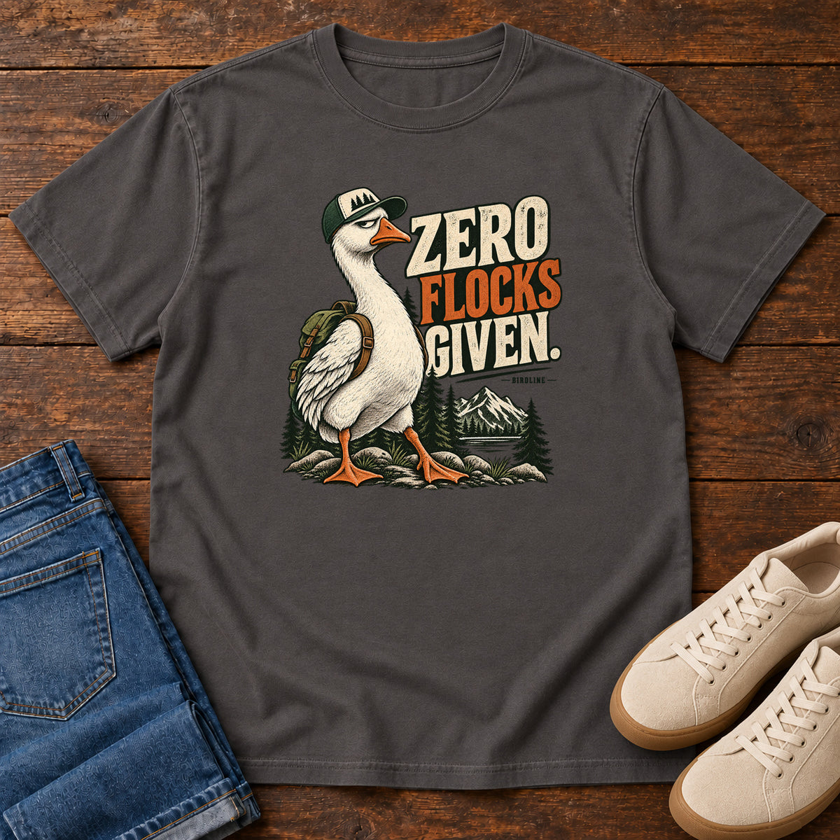 Zero Flocks Given T-Shirt in Graphite, front laid flat on wood, featuring a white long-necked bird wearing a cap and backpack beside cream-and-orange lettering and mountains.