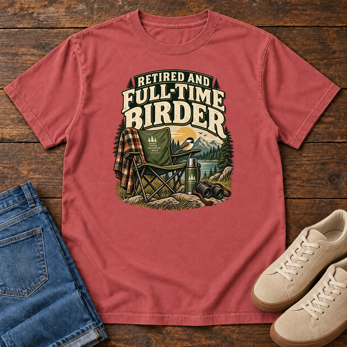 Crimson Retired and Full-Time Birder T-Shirt with a camp chair, plaid shirt, flask and binoculars in a mountain-lake scene, front flat lay on wood.
