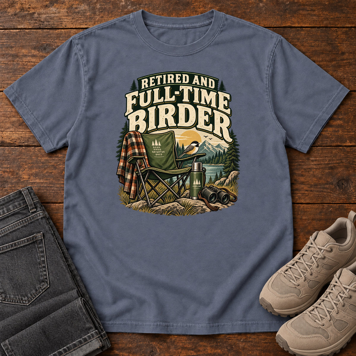 Denim Retired and Full-Time Birder T-Shirt with a camp chair, plaid shirt, flask and binoculars in a mountain-lake scene, front flat lay on wood.