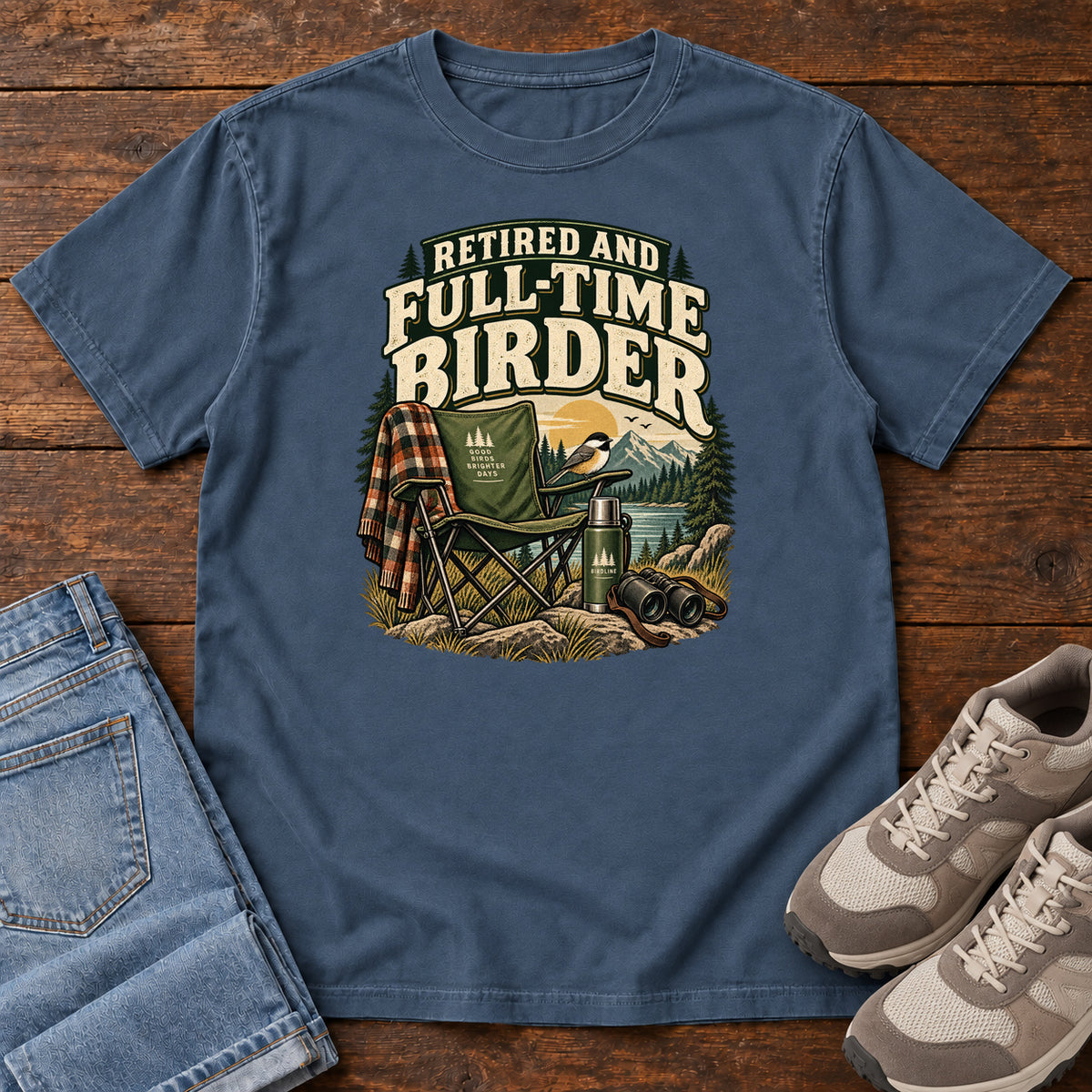 Midnight Retired and Full-Time Birder T-Shirt with a camp chair, plaid shirt, flask and binoculars in a mountain-lake scene, front flat lay on wood.