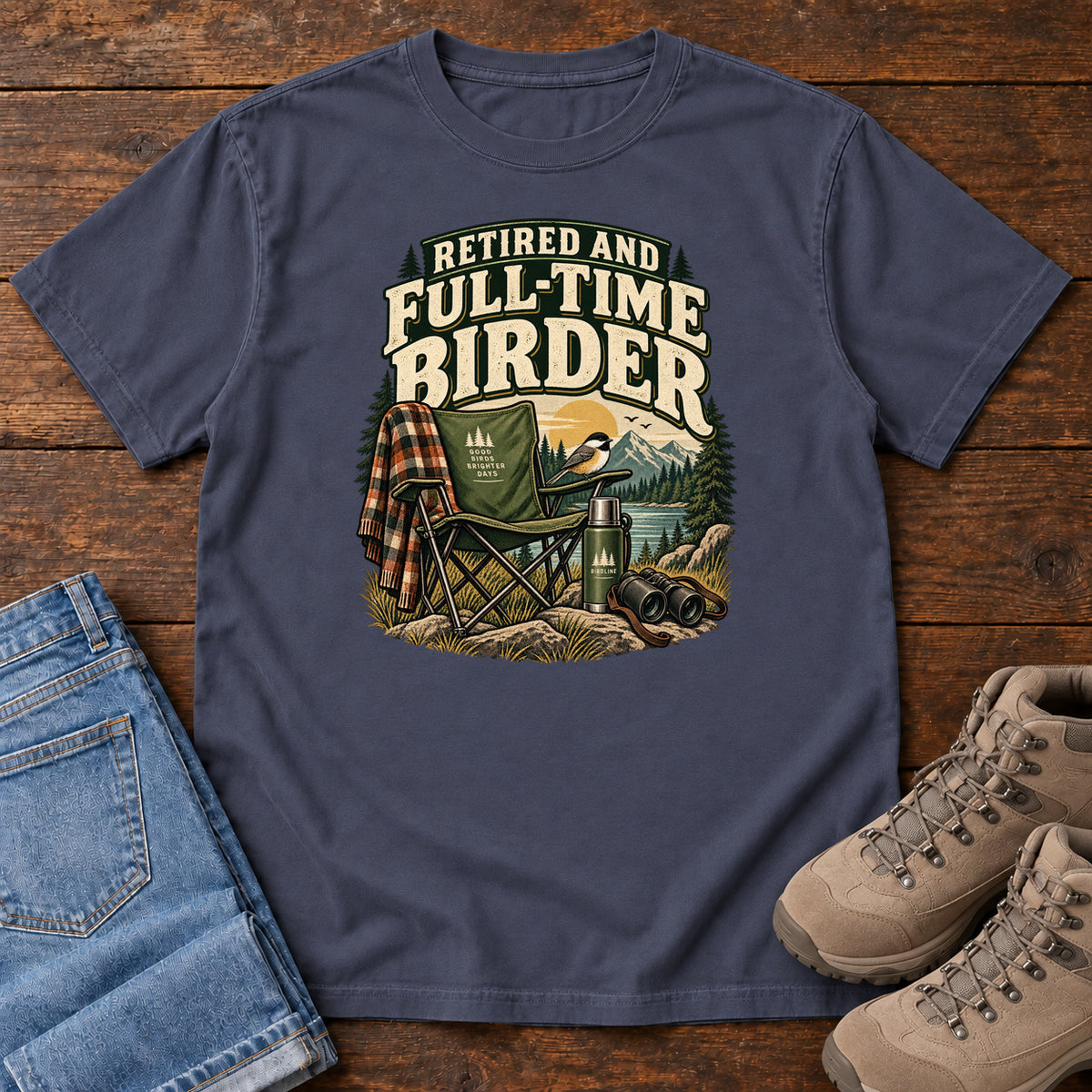 Navy Retired and Full-Time Birder T-Shirt with a camp chair, plaid shirt, flask and binoculars in a mountain-lake scene, front flat lay on wood.