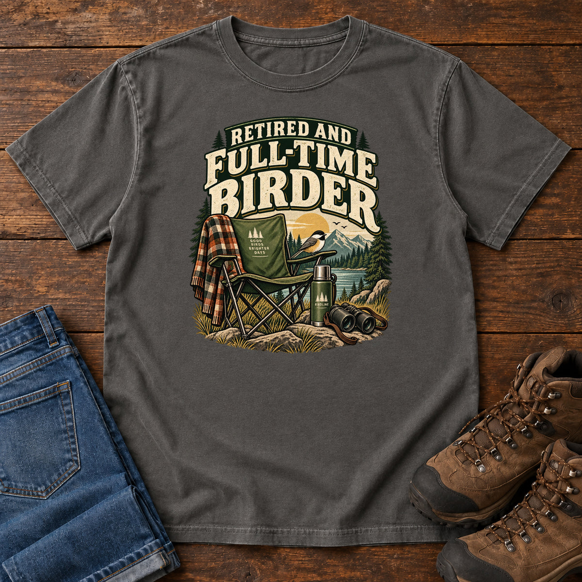 Pepper Retired and Full-Time Birder T-Shirt with a camp chair, plaid shirt, flask and binoculars in a mountain-lake scene, front flat lay on wood.