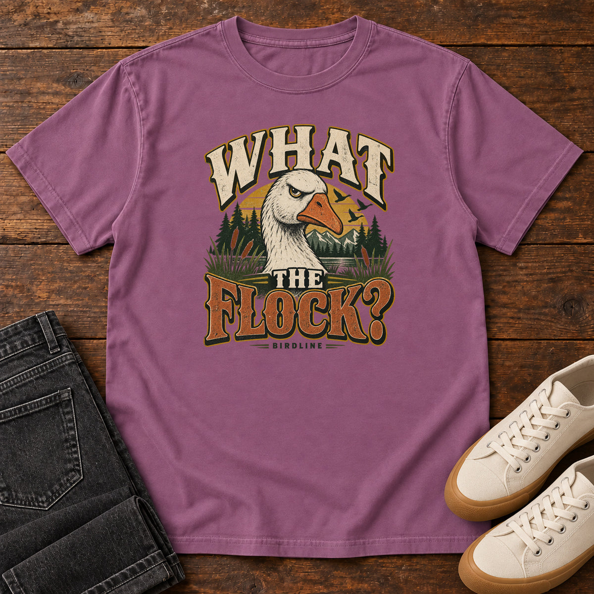 Berry What the Flock? T-Shirt with a white goose head, evergreens and cream and orange slogan lettering; front flat lay on wood.