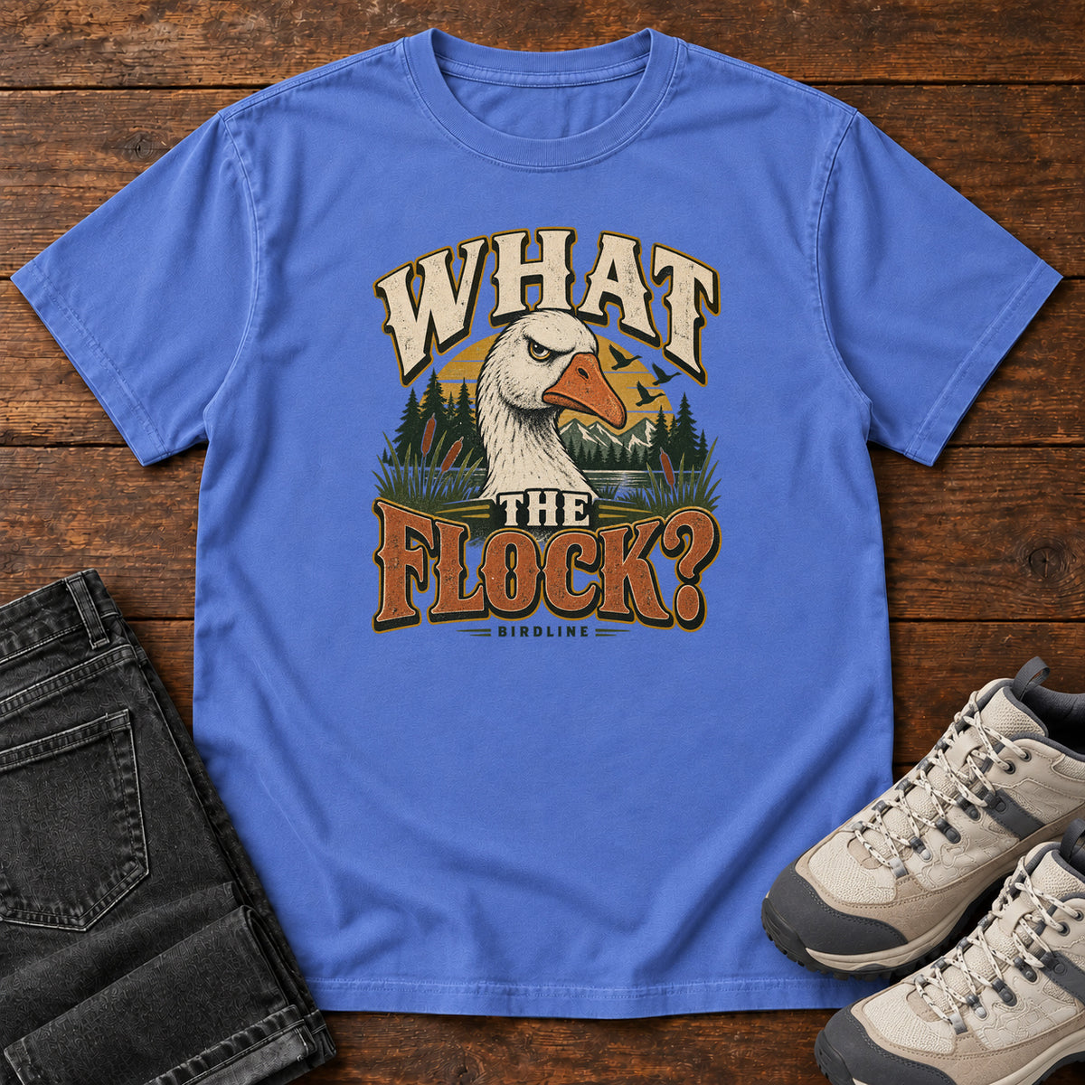 Mystic Blue What the Flock? T-Shirt with a white goose head, evergreens and cream and orange slogan lettering; front flat lay on wood.